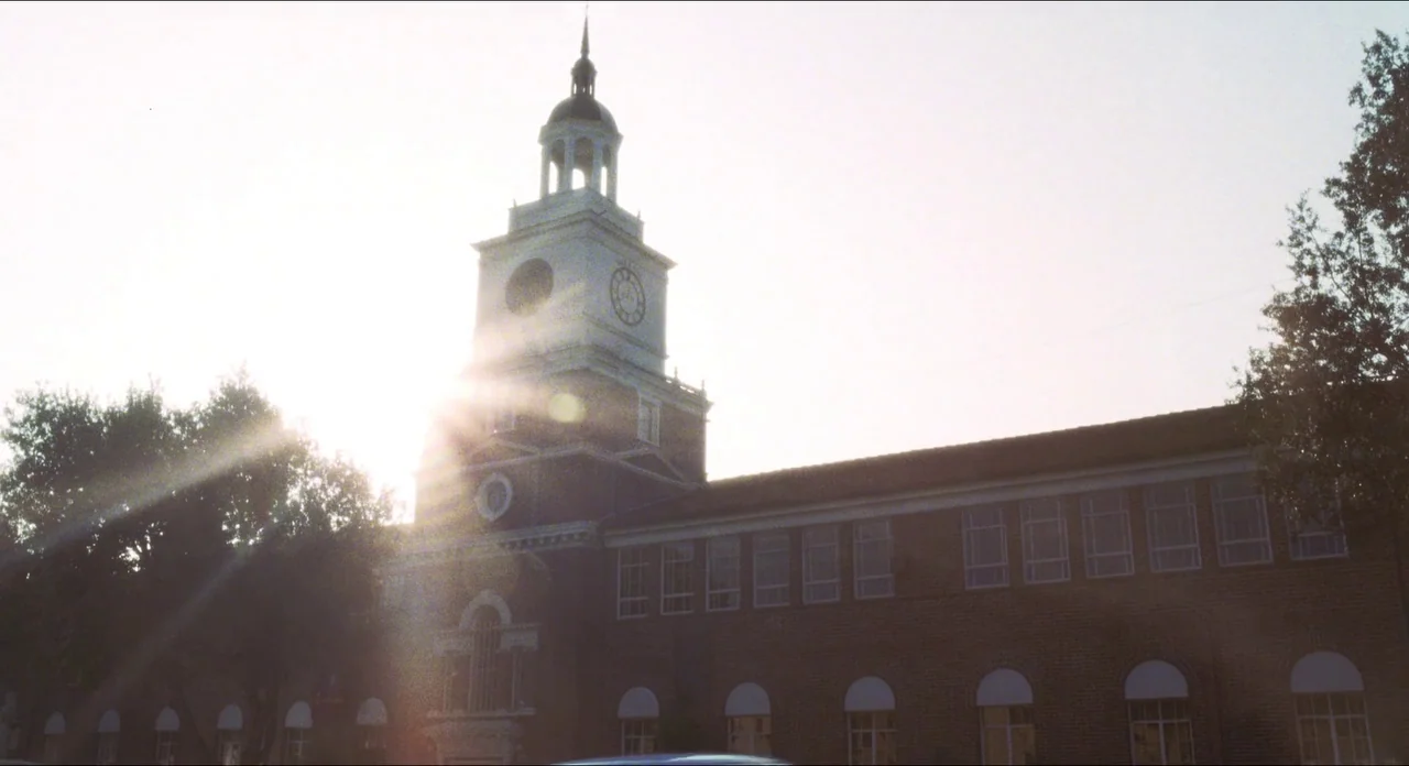 Catch Me If You Can (2002) movie still: wide — A brick school building features a central clock tower with a prominent cupola. The sun sits directly…