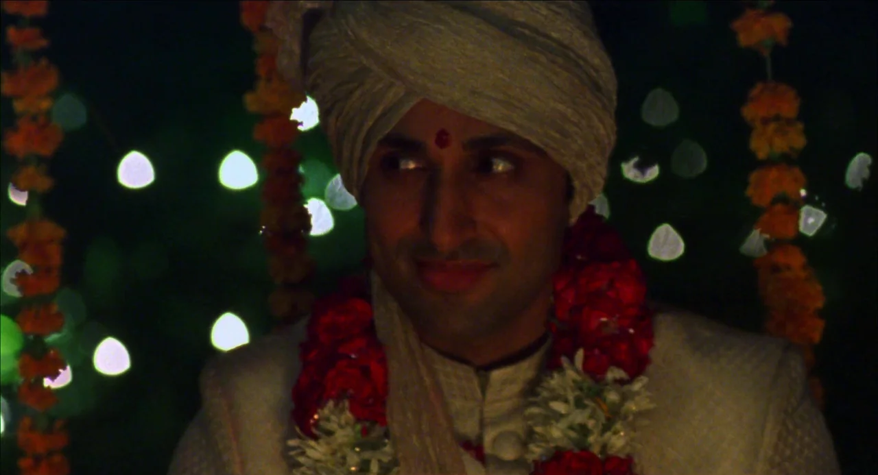 Monsoon Wedding (2001) movie still: closeup — A man wearing traditional wedding attire sits in a dark, festive interior space. Garlands of flowers…