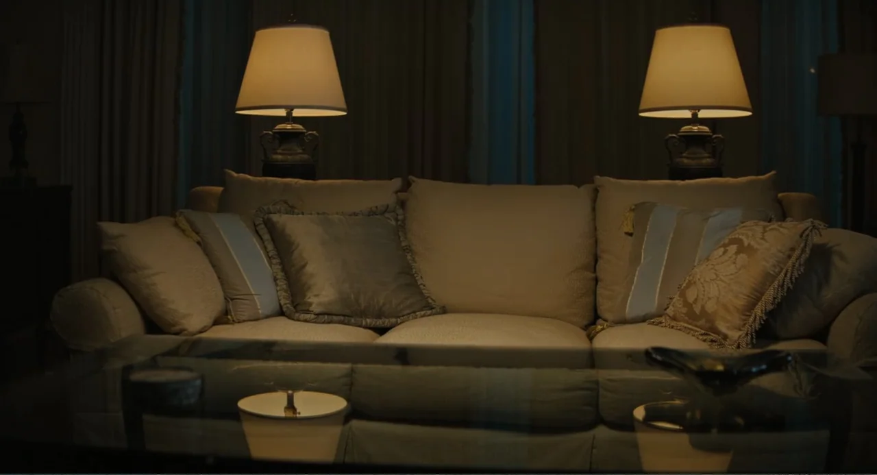 Beautiful Boy (2011) movie still: medium — A dimly lit room features a plush upholstered sofa against a dark wall. Two lamps cast warm circles of…
