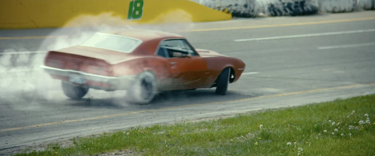 God Bless the Broken Road (2018) movie still: medium — A worn asphalt racetrack is lined with a yellow concrete barrier and grassy verge. An orange…