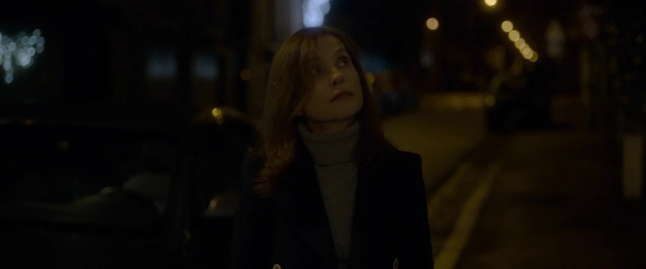 Elle (2016) movie still: medium — A dark city street at night features a woman standing on the sidewalk. She is framed from the waist up as she…