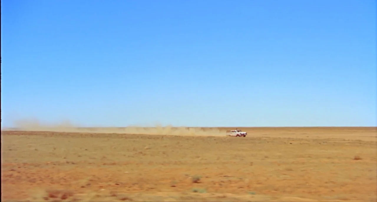 Wake In Fright (1971) movie still: establishing — A vast, flat desert landscape extends to a low, distant horizon under a sky. A small white vehicle…