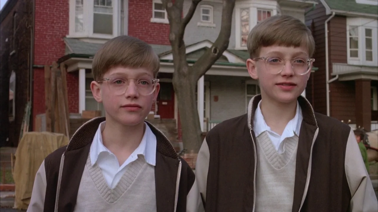 Dead Ringers (1988) movie still: medium — Two identical young boys stand in a suburban residential neighborhood during the day. Houses with brick…