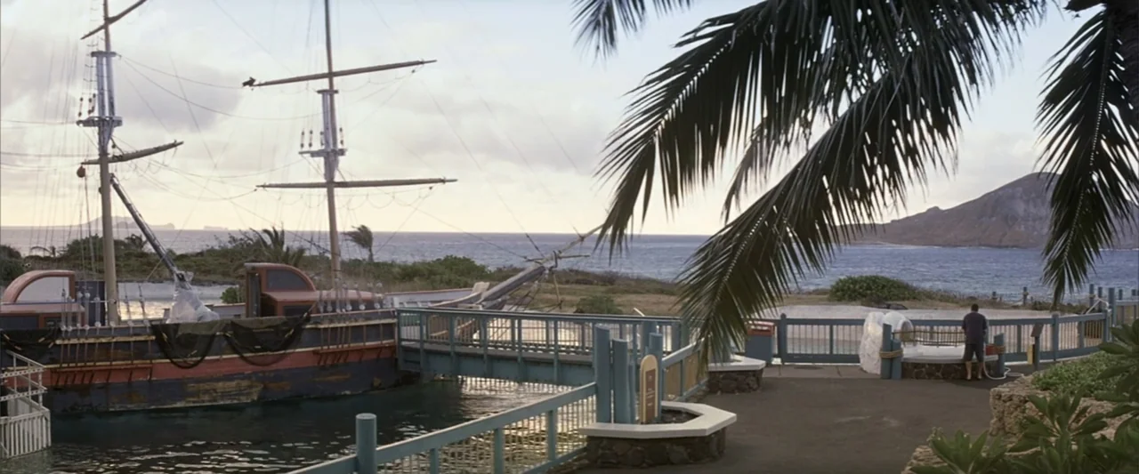50 First Dates (2004) movie still: wide — A wooden ship is docked at a pier overlooking a coastal tropical landscape. A lone man stands on the pier…