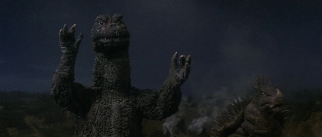 Godzilla Vs Gigan (1972) movie still: medium — Two giant monsters stand in a dark, sparse landscape during the night. The creatures face toward the…