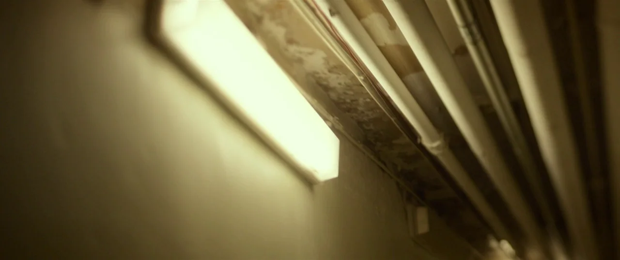 Split (2016) movie still: closeup — A weathered industrial wall features an illuminated fluorescent light fixture with exposed overhead pipes. The…