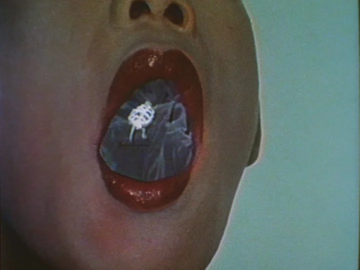 Les Chants de Maldoror (1977) movie still: closeup — An extreme close-up of an open human mouth against a solid pastel background. A small white…