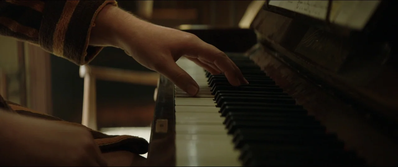 Rocketman (2019) movie still: closeup — An interior space features an upright piano with vintage ivory keys. A person wearing a striped robe rests…
