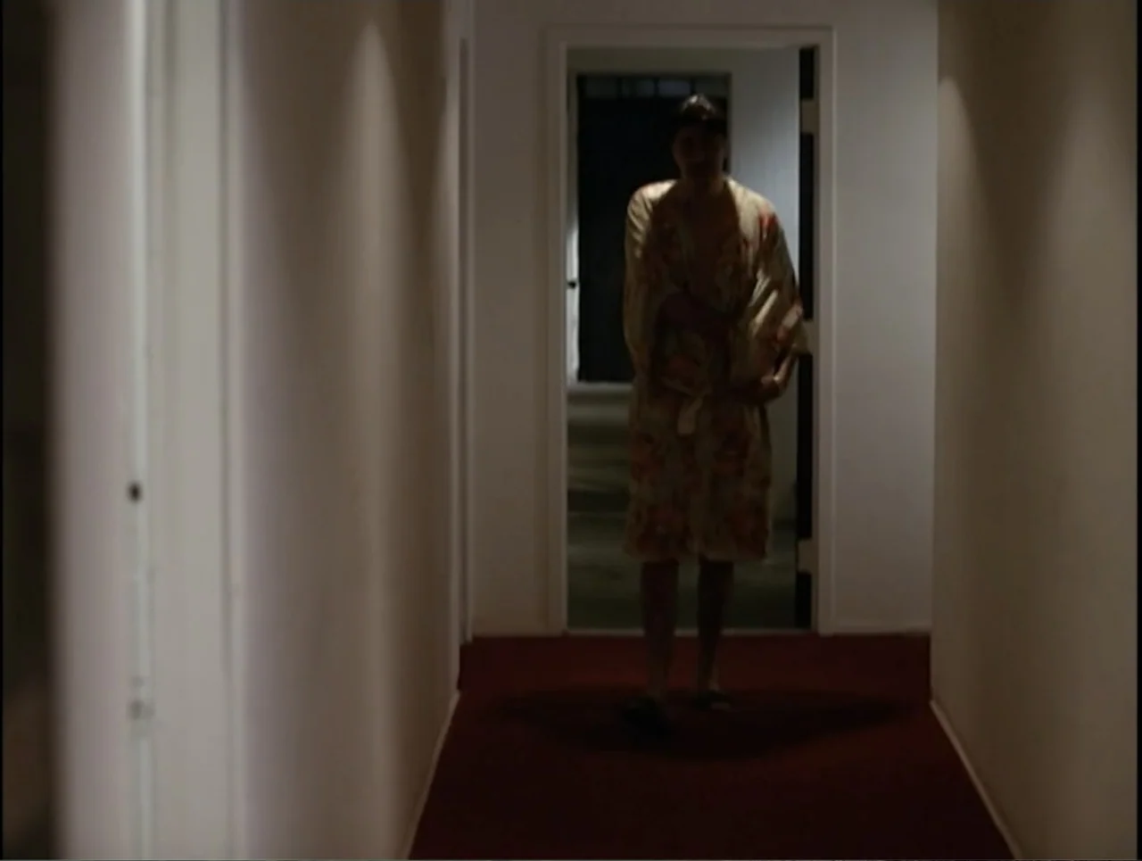 Apartment 12 (2001) movie still: fullbody — A dimly lit narrow hotel corridor features white walls and a dark red carpet. A man wearing a patterned…