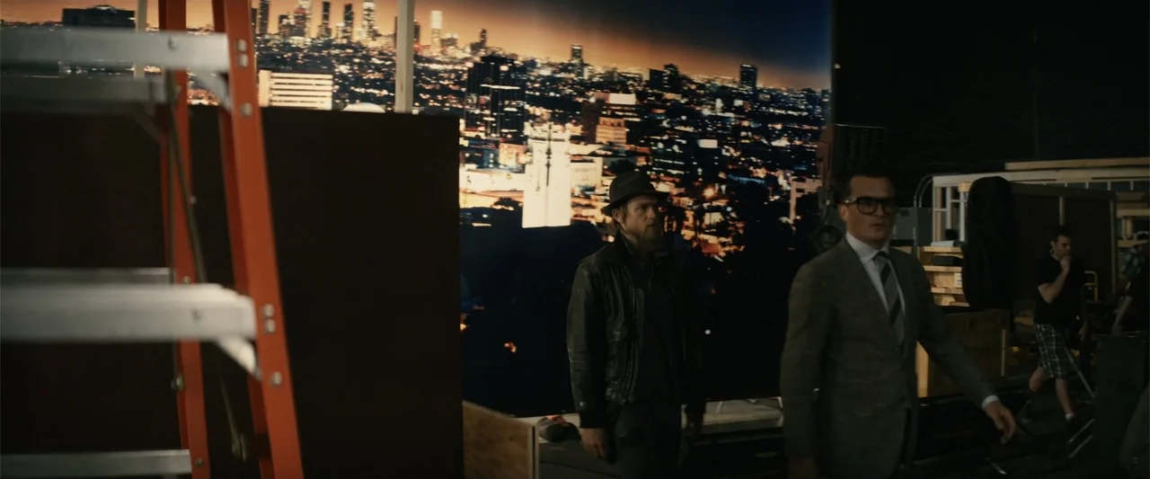 Last Looks (2022) movie still: medium — A cluttered interior film studio features a massive, illuminated city night mural background. Two men in…