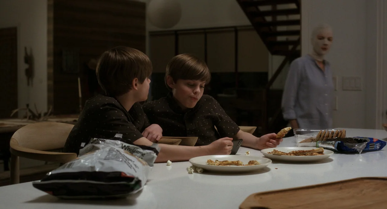 Goodnight Mommy (2022) movie still: medium — Two young boys eat snacks at a white kitchen island in a contemporary home. A woman with her head fully…