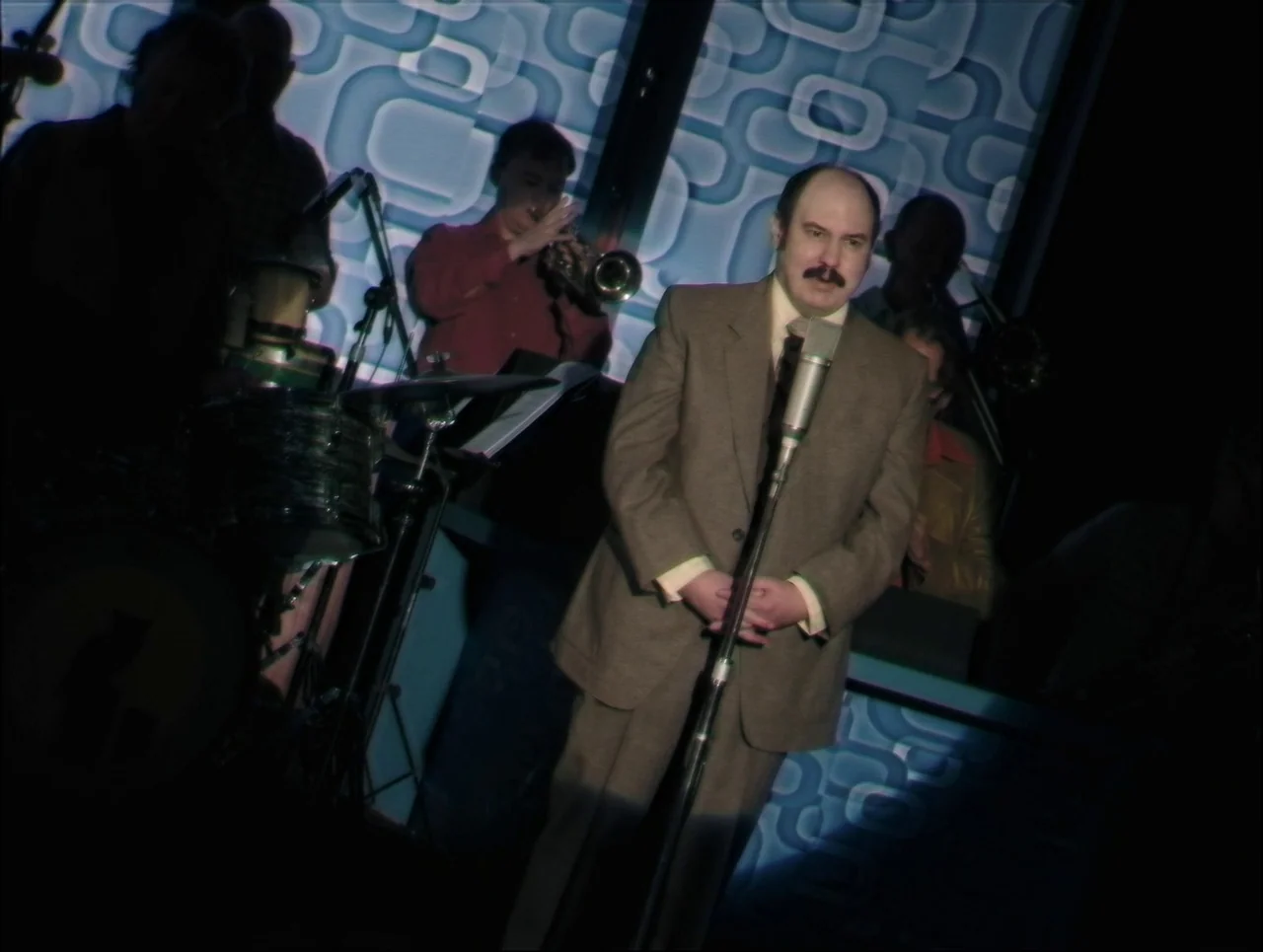 Late Night With The Devil (2023) movie still: medium — A dimly lit club features a retro blue geometric pattern backdrop behind a band. A man in a…