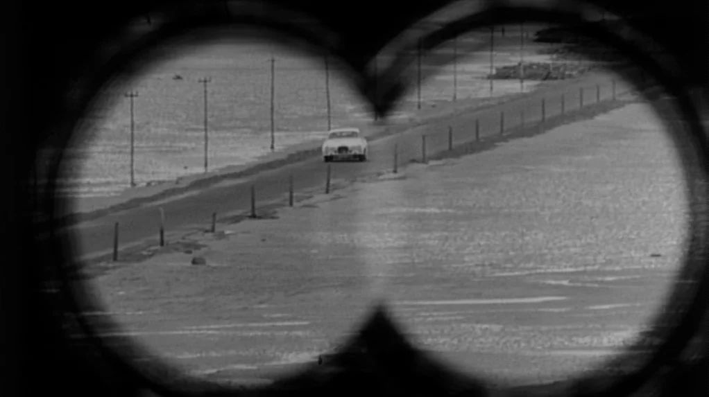 Cul-de-sac (1966) movie still: wide — A flat, desolate coastal road stretches through a barren landscape under an overcast sky. A single vintage…