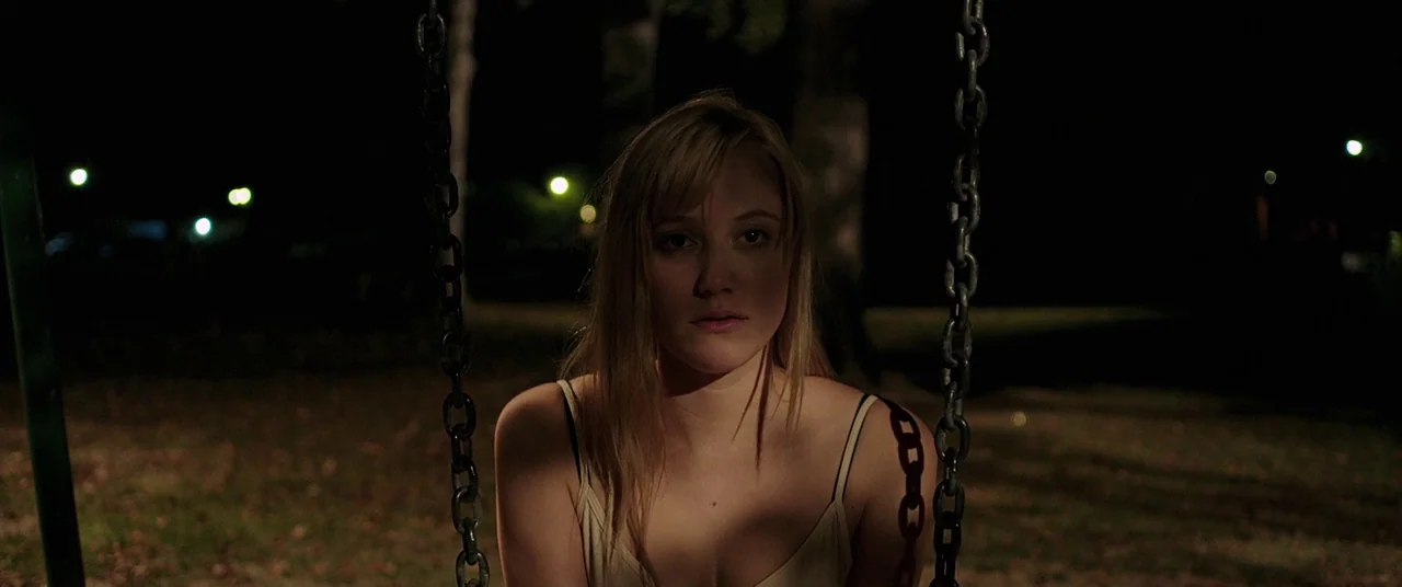 It Follows (2015) movie still: medium — A night scene at a playground with distant blurred city lights visible. A young woman sits on a swing in the…