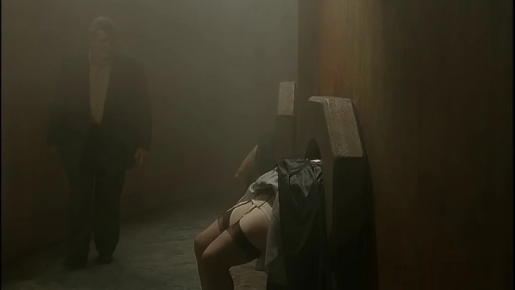 Romance (1999) movie still: medium — A dimly lit, hazy stone cell features a heavy wooden pillory against a wall. A man in a dark suit stands nearby…