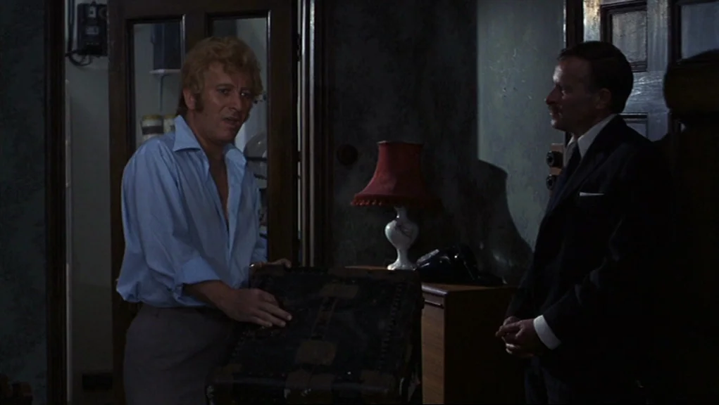 Frenzy (1972) movie still: medium — A dimly lit room with dark wallpaper and a glass-paneled wooden door in back. A man with blond hair holds a…