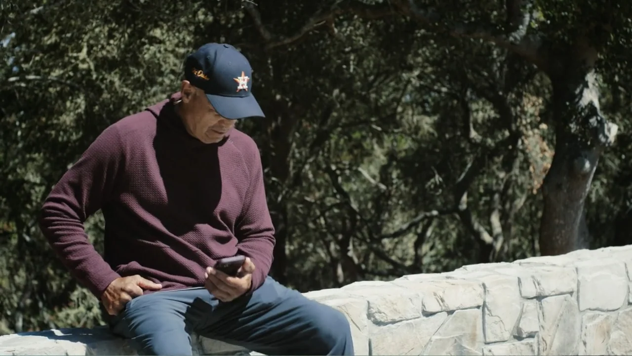Reggie (2023) movie still: medium — An outdoor area features a light-colored stone wall in front of dark trees. A man wearing a baseball cap and…