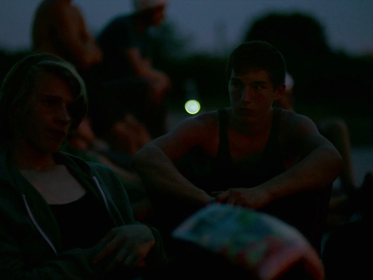 Violet (2014) movie still: medium — Two young men sit together outdoors at night during an evening gathering. Other figures are obscured in the dark…