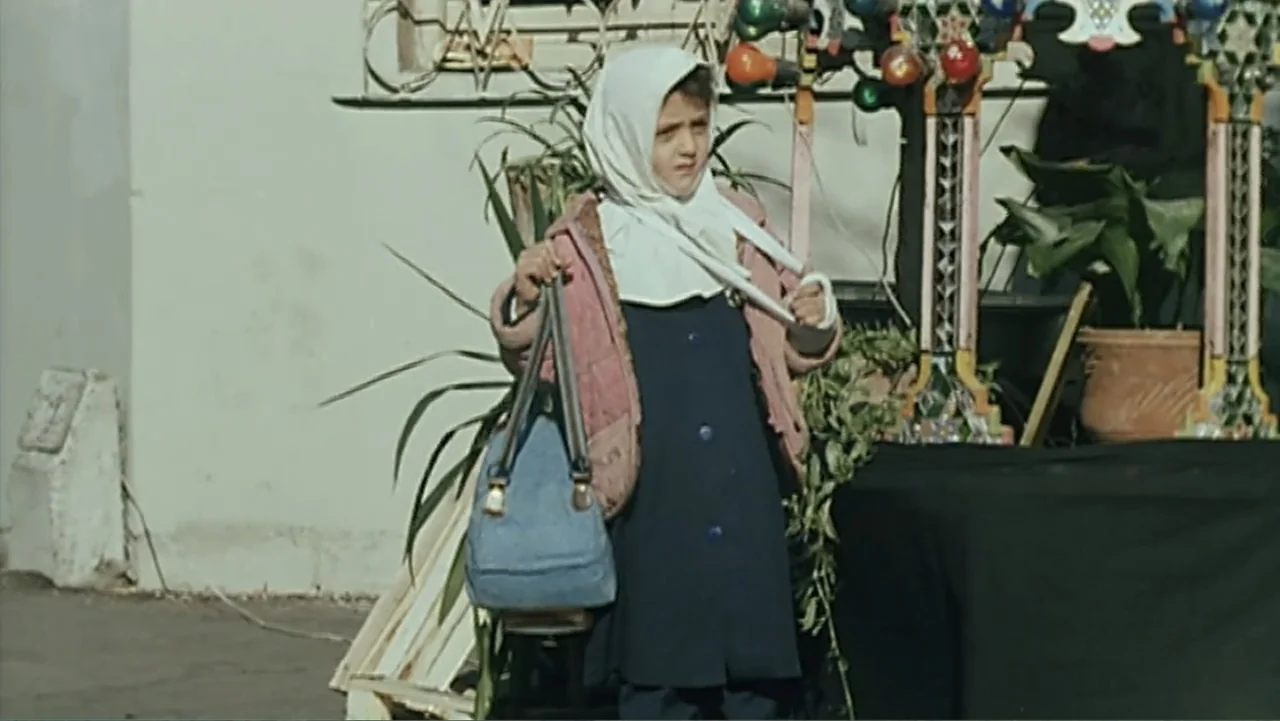 The Mirror (2014) movie still: medium — A young girl stands on a paved area near a wall and potted greenery. She wears a headscarf and a coat while…