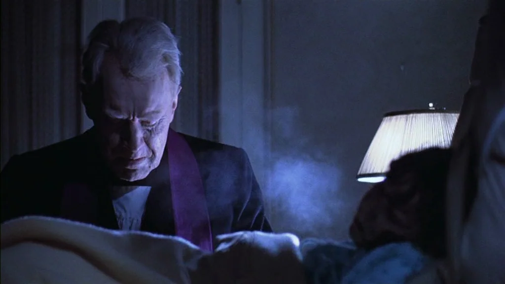 The Exorcist (1973) movie still: medium — An interior bedroom at night features a priest standing over a prone, covered figure. Cool blue mist rises…
