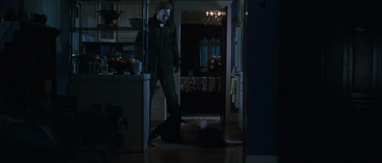 Halloween (2007) movie still: fullbody — A dark residential interior features a person lying prone on a hardwood floor. A masked man stands directly…