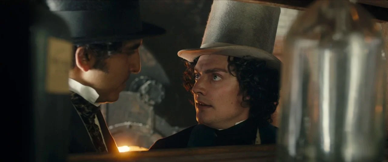 The Personal History of David Copperfield (2019) movie still: medium — Two men wearing 19th-century attire converse in a low-light space filled with…