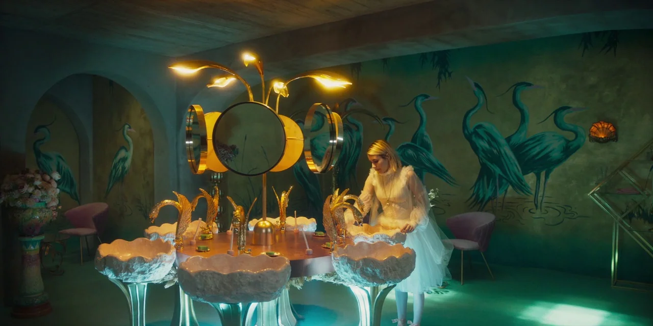 Paradise Hills (2019) movie still: wide — An opulent vanity room features walls covered in a teal crane mural. A woman in a white dress stands at a…