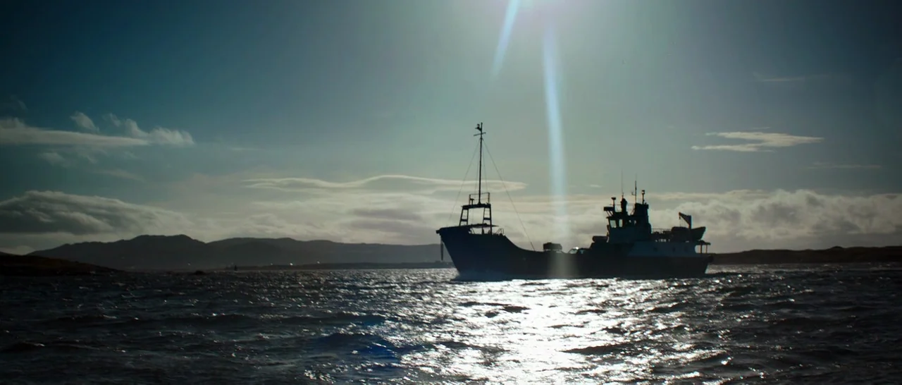 The Hallow (2015) movie still: wide — A fishing boat moves across choppy water under a bright sun and cloudy sky. Distant hills line the horizon in…