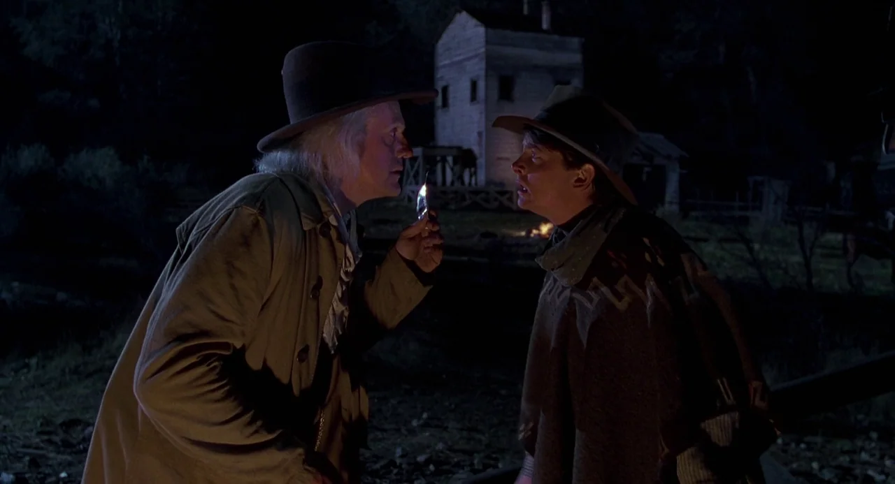 Back To The Future Part III (1990) movie still: medium — A dimly lit rural location with a wooden structure in the background at night. Two men in…