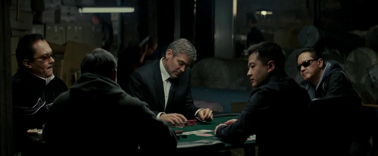 Michael Clayton (2007) movie still: medium — A cluttered, dimly lit industrial space features boxes and rolls of material in shadows. Five men sit…