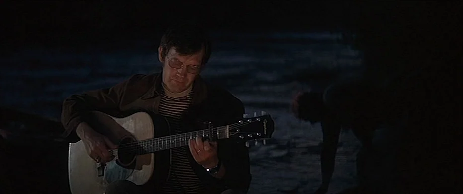 Deliverance (1972) movie still: medium — A man sits on a dark riverbank at night with a guitar. He focuses on playing the instrument while another…