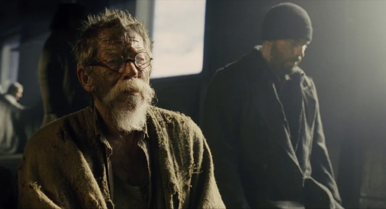 Snowpiercer (2013) movie still: medium — A dark, dilapidated indoor space features a large window with muted daylight streaming through. Two men sit…