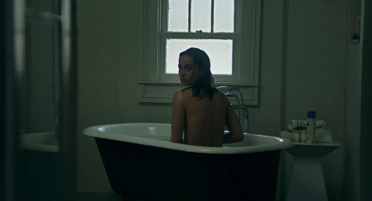 Deep Water (2022) movie still: medium — A dark, dimly lit bathroom features a black-and-white clawfoot tub near a window. A woman with wet hair sits…