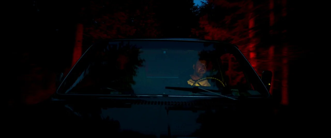 Mandy (2018) movie still: medium — A dark vehicle travels along a road flanked by blurred, red-lit trees at night. Two men occupy the front seats of…