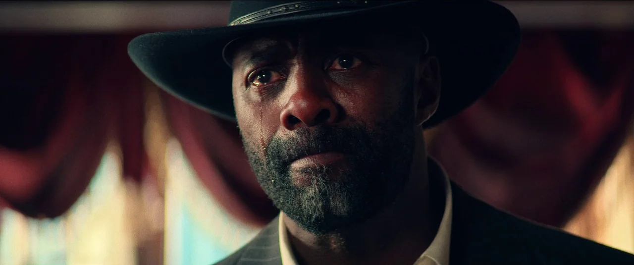The Harder They Fall (2021) movie still: closeup — A man stands before blurry deep red curtains inside a dimly lit room. He wears a dark cowboy hat…