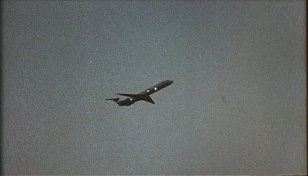 Labyrinth of Passion (1982) movie still: wide — An airplane traverses a clear, monochromatic sky during the daytime. The aircraft is captured from a…