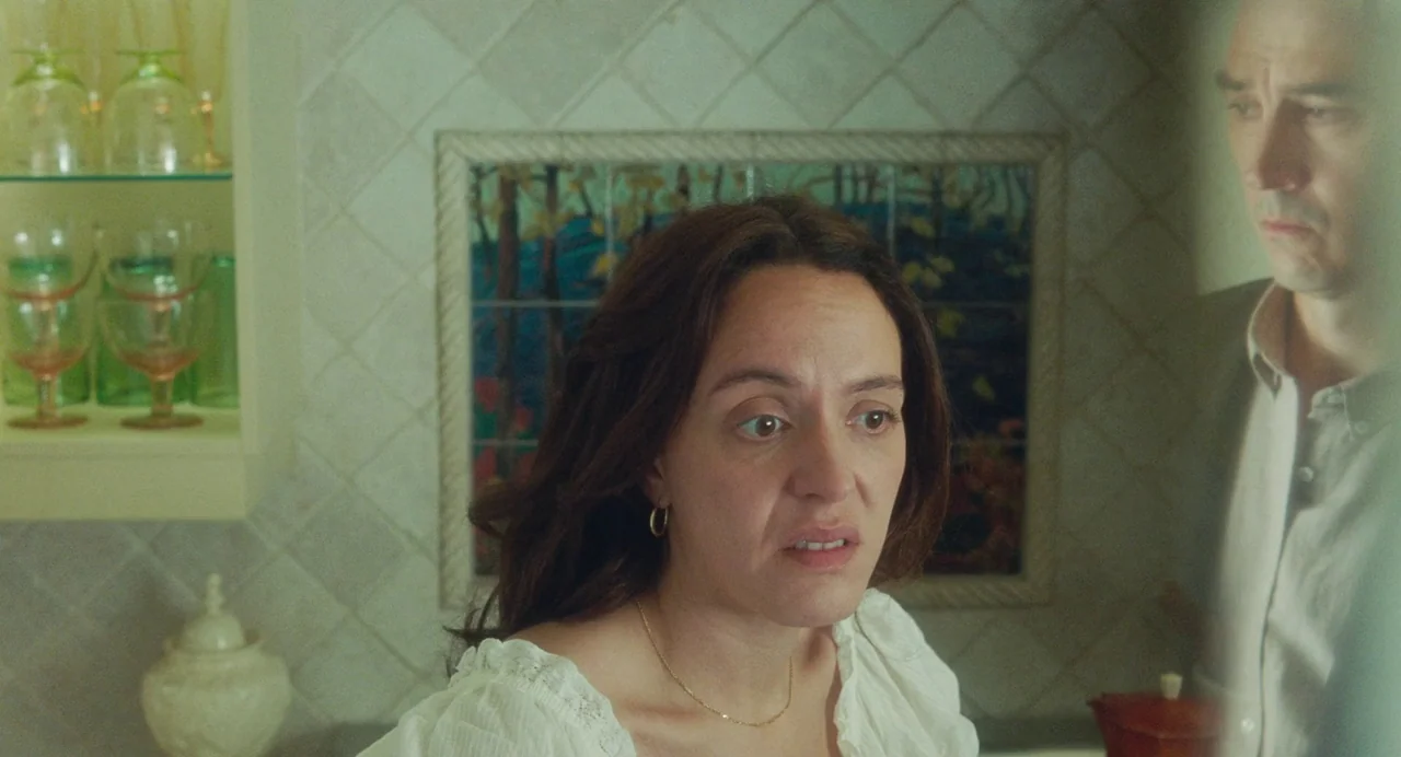 Babysitter (2022) movie still: medium — A residential kitchen with light green tiled walls and decorative forest-themed tiles. A woman in a white…