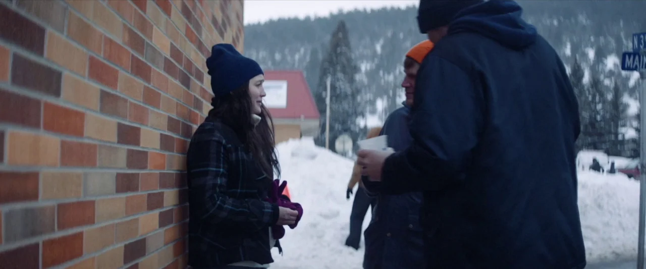 The Unknown Country (2022) movie still: medium — A sidewalk next to a brick building features heavy snow banks along the road. A woman leans against…