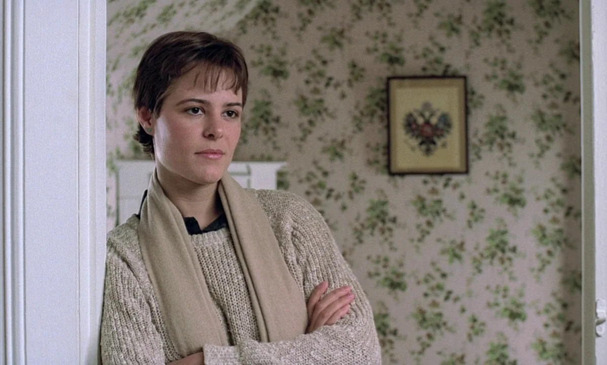 A Tale of Springtime (1990) movie still: medium — A room with floral wallpaper features a framed double-headed eagle emblem on the wall. A young…