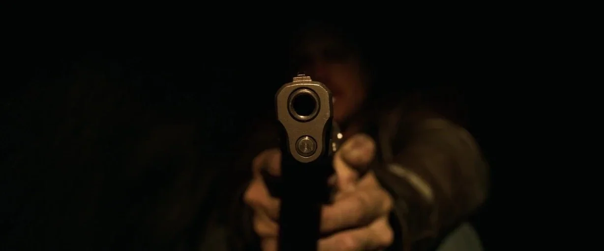 Breaking Bad (2008) movie still: closeup — A person stands in a dimly lit, dark space holding a firearm forward. The barrel of the gun is in sharp…