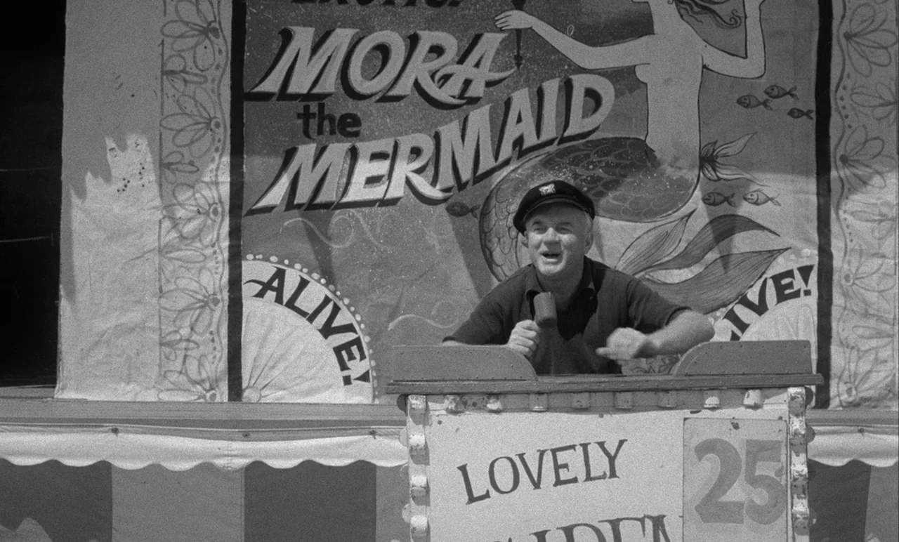 Night Tide (1961) movie still: medium — A carnival barker stands behind a wooden counter advertising a mermaid sideshow attraction. Behind him hangs…