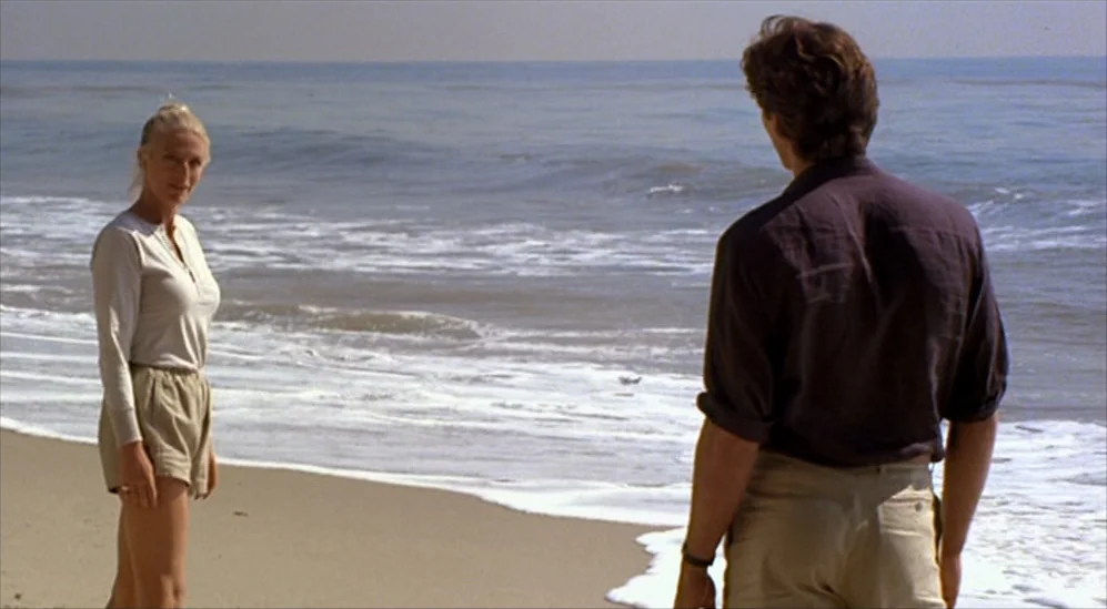 American Gigolo (1980) movie still: fullbody — A flat sandy beach stretches toward the ocean under an overcast sky. A woman stands on the left…