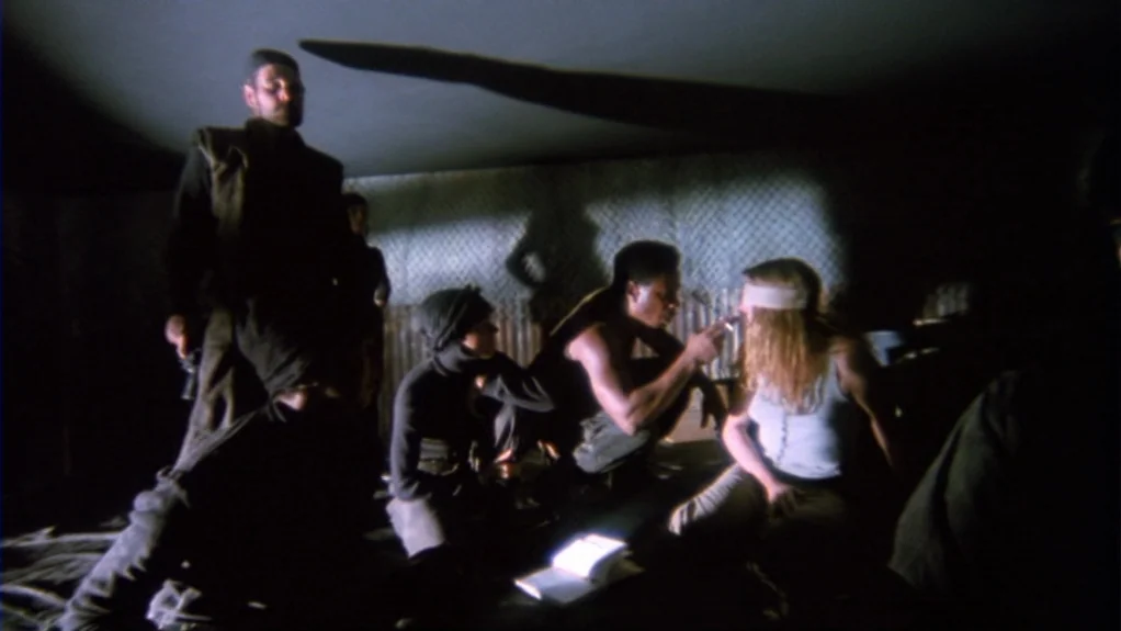 Patty Hearst (1988) movie still: medium — A dimly lit, cramped makeshift shelter interior features walls of corrugated metal or fencing. A small…