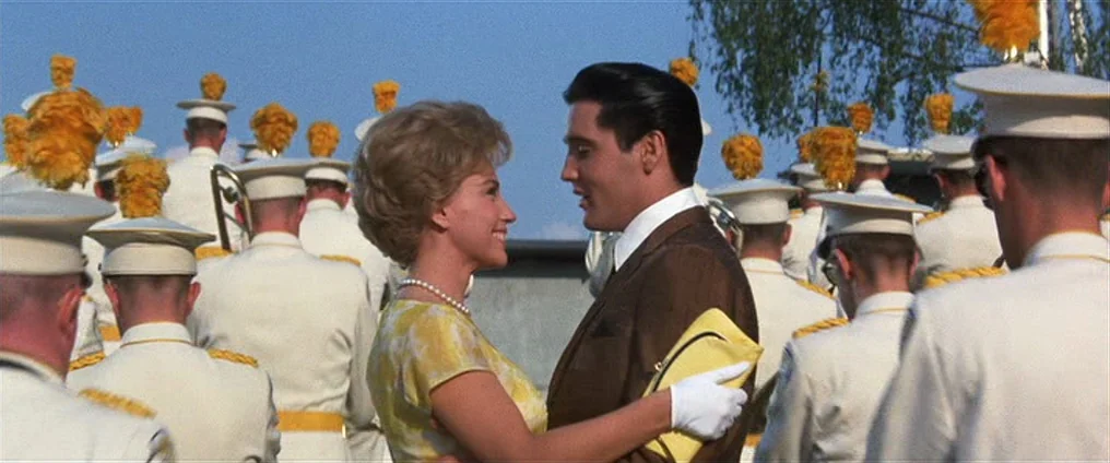 It Happened At The World’s Fair (1963) movie still: medium — A man and woman stand together in a crowd of soldiers in dress uniforms. The soldiers…