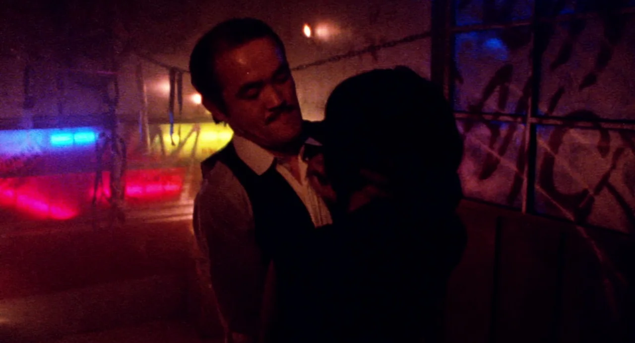 Burst City (1982) movie still: medium — A dark space features a metal frame wall with graffiti and saturated neon lights. A man in a waistcoat…