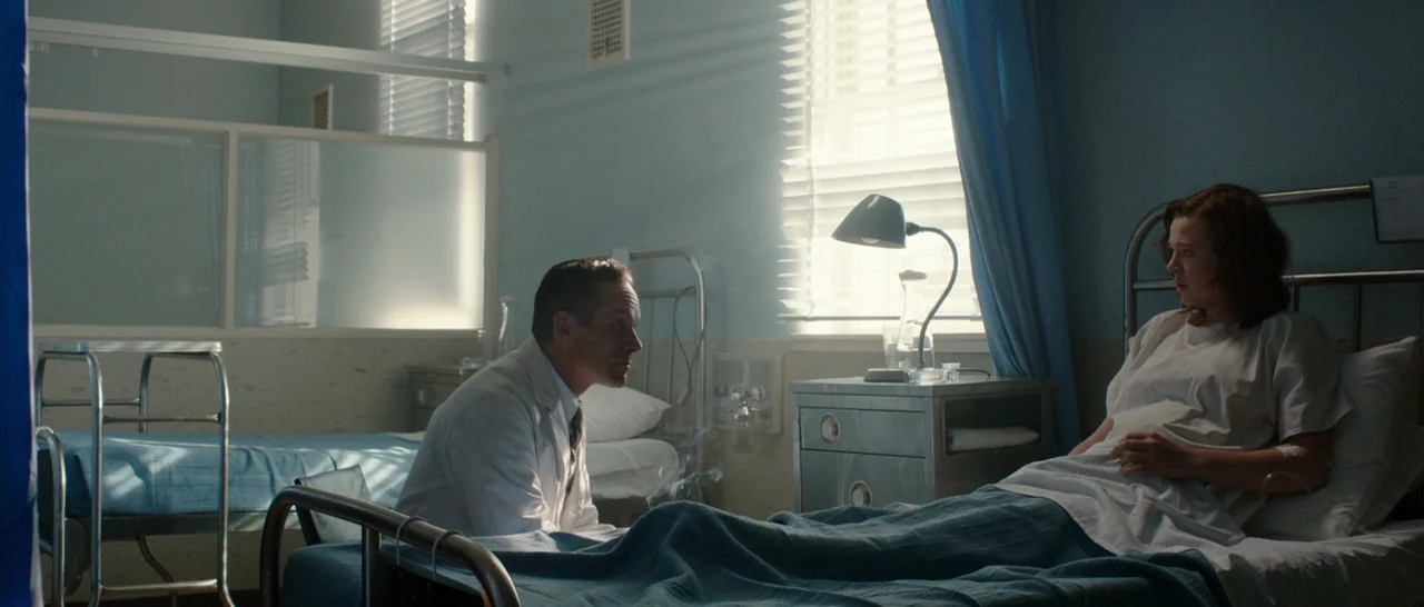 Predestination (2014) movie still: wide — A sterile medical space with blue walls and metal furniture lit by window sunlight. A doctor in a white…