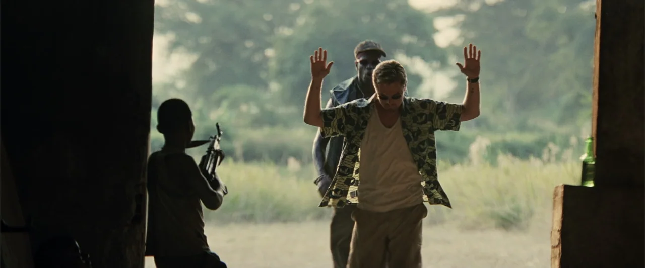 Blood Diamond (2006) movie still: medium — A dilapidated building interior opens onto an overgrown field during the day. A man with hands raised is…