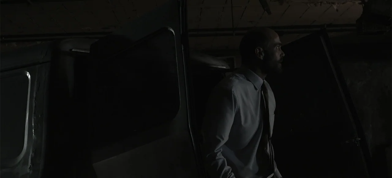 About the Deaths (2016) movie still: medium — A dimly lit garage interior features a parked vehicle with an open side door. A man in a dress shirt…