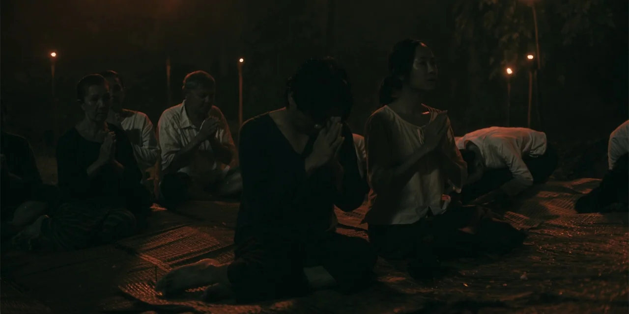 Host (2020) movie still: medium — An outdoor ground covered with woven mats at night illuminated by distant torches. A group of men and women sit on…