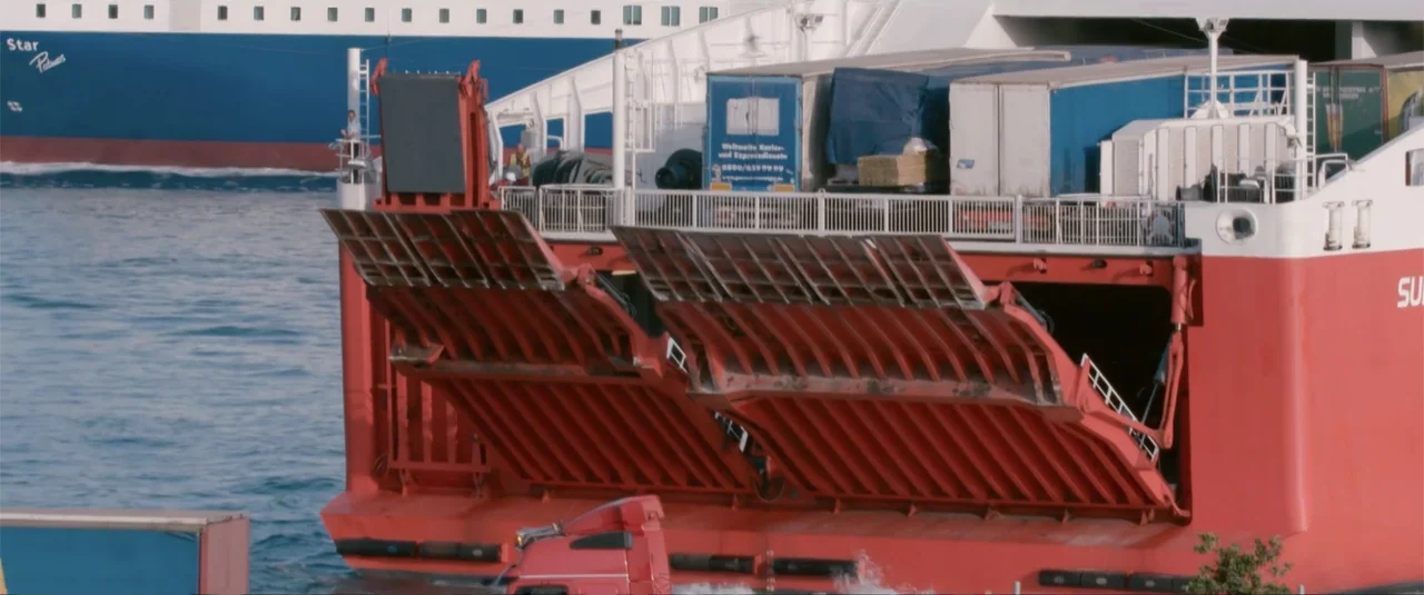 Simple as Water (2021) movie still: medium — A large red ferry with its loading ramp lowered sits on the water. Another blue and white ship is…